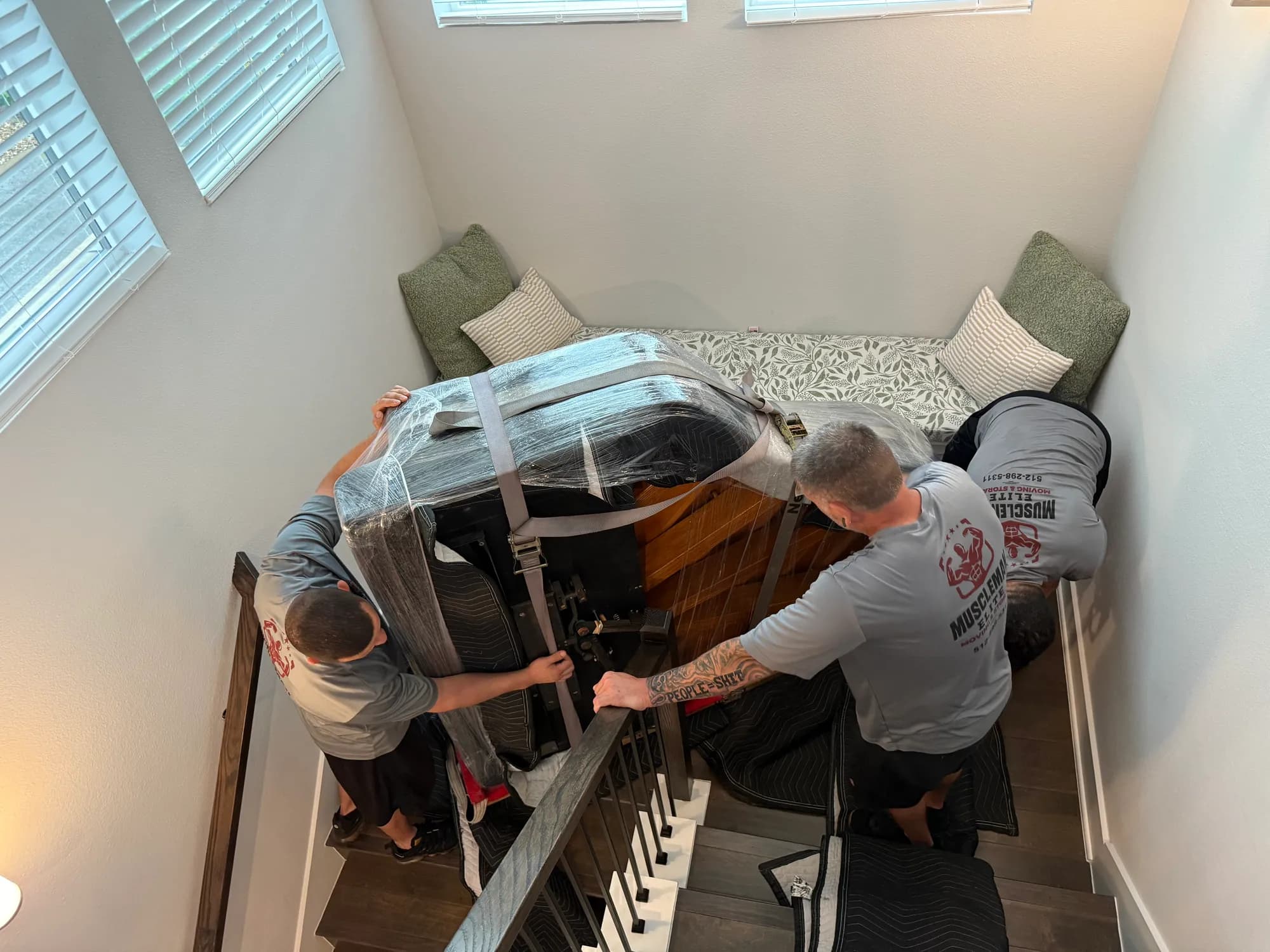 Muscleman Elite crew easing a blanket-wrapped piano down a staircase on boards