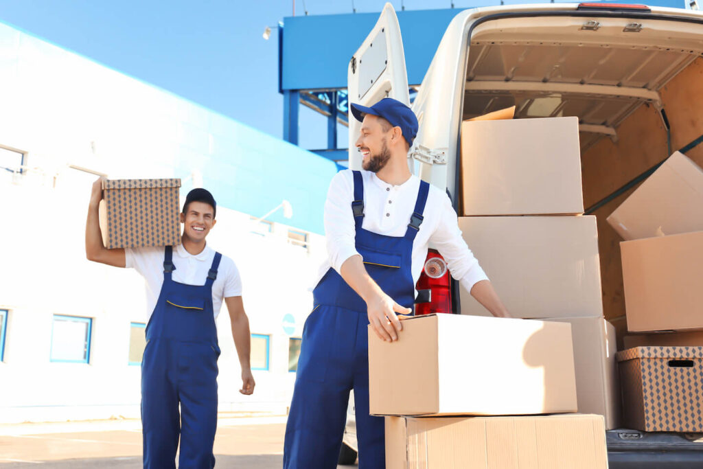 Two movers wearing white shirts and blue coveralls taking boxes out of the back of a moving truck.