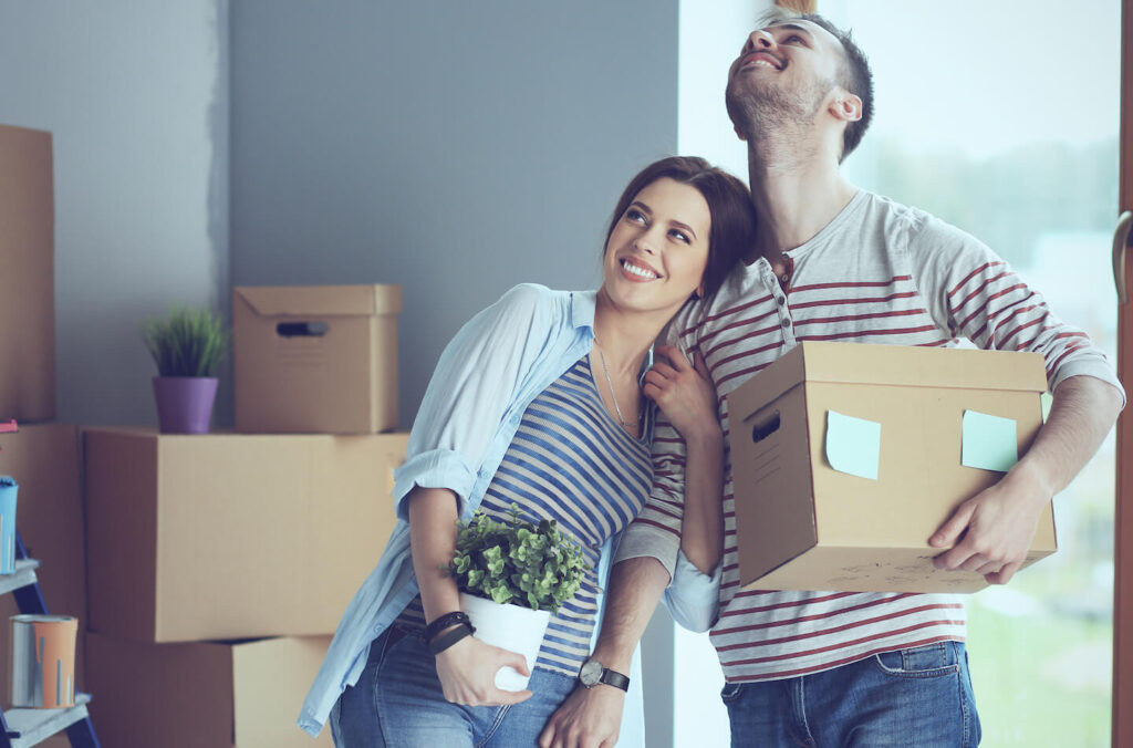 A white woman holding a house plant smiling and leaning her head against a white man's shoulder; the man is holding a cardboard box. Both are looking up toward the ceiling in a room filled with moving boxes.